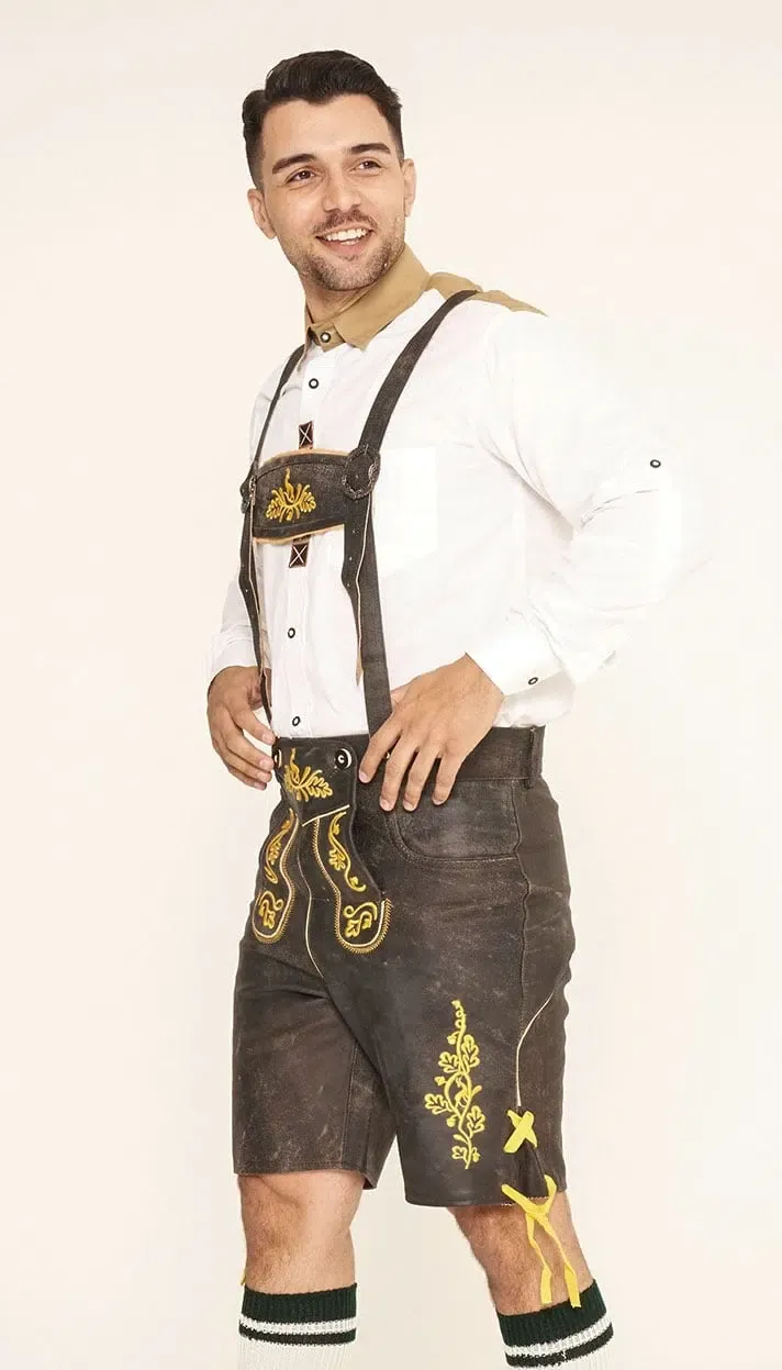 Side view of a man posing in dark brown Lederhosen with ornate yellow embroidery, matching suspenders, and a white button-down shirt. - Lederhosen For Sale