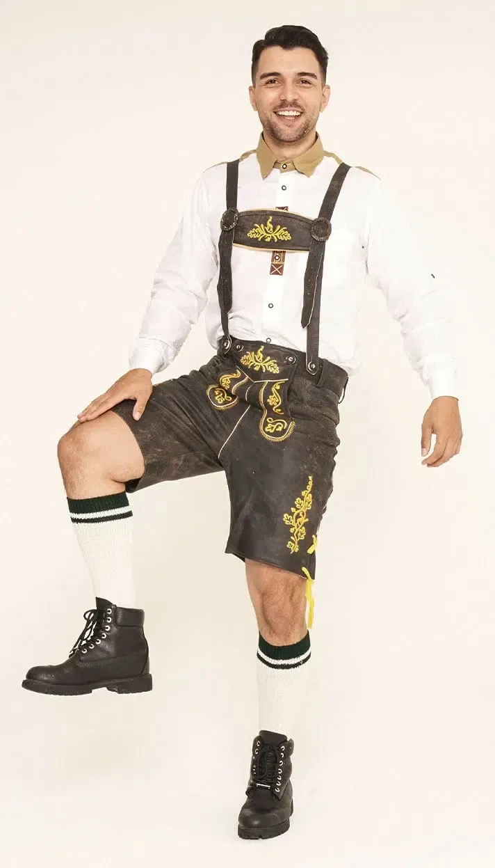 Smiling man posing in dark brown Lederhosen with yellow embroidery, a white shirt, white socks with green stripes, and black boots. - Lederhosen For Sale