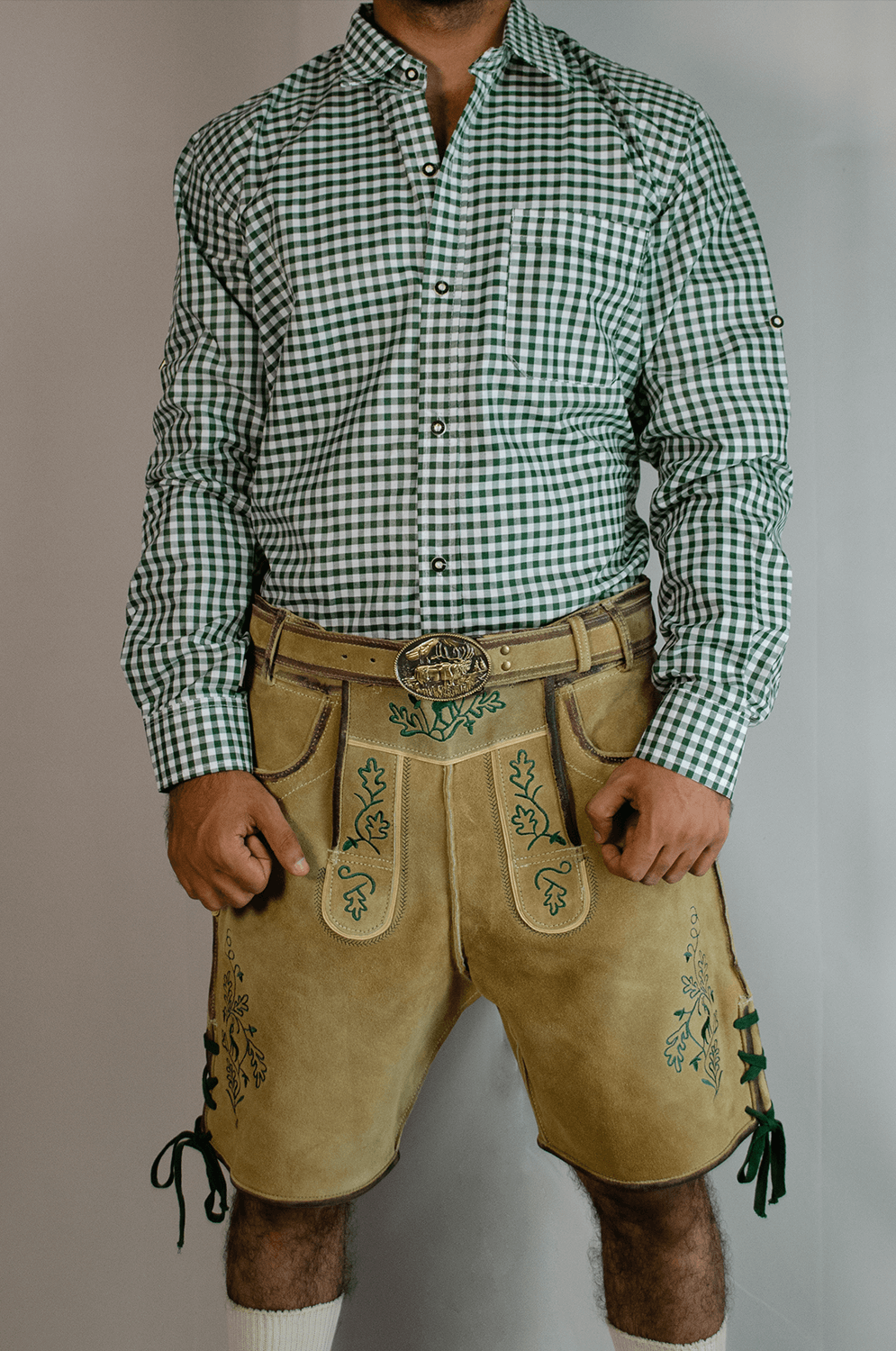 Front view of a man wearing light gold handcrafted Lederhosen shorts with a large gold belt buckle, paired with a green and white checkered long-sleeve shirt. - Lederhosen For Sale
