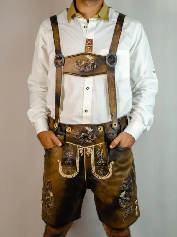 Front view of a man standing with hands in pockets wearing dark chocolate distressed leather Lederhosen with matching stag-embroidered suspenders and a white shirt. - Lederhosen For Sale