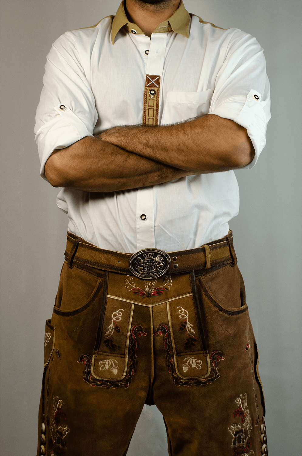 Front view of a man standing with arms crossed wearing vintage brown embroidered Lederhosen with a matching leather belt and a white shirt. - Lederhosen For Sale