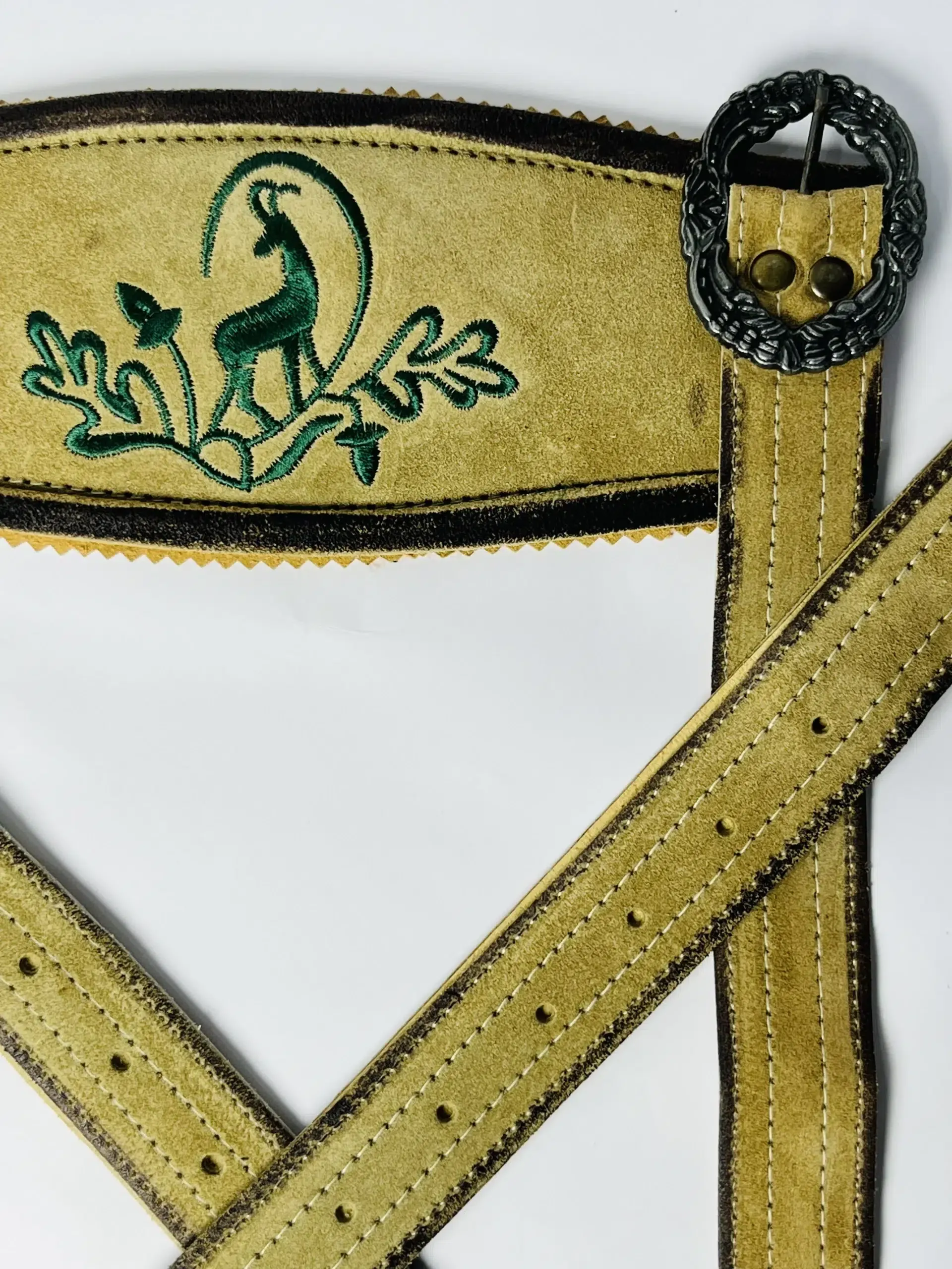 Close-up of green stag and oak leaf embroidery on the front cross-bridge of light gold Lederhosen suede suspenders with a detailed dark silver metal buckle. - Lederhosen For Sale