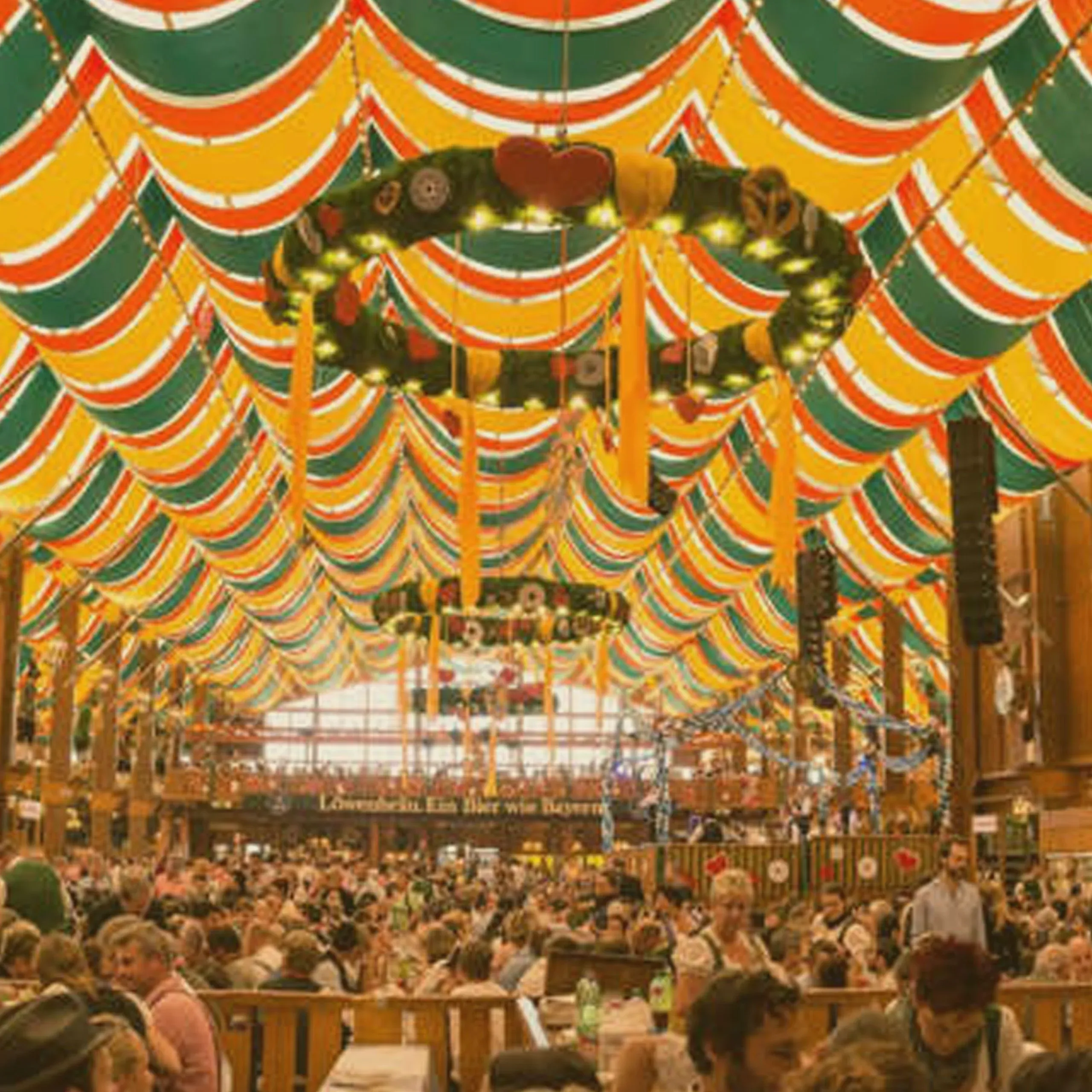 Traditional Oktoberfest beer tent crowded with people, featuring green, orange, and yellow draped fabric ceilings and large decorative wreaths. - Lederhosen For Sale