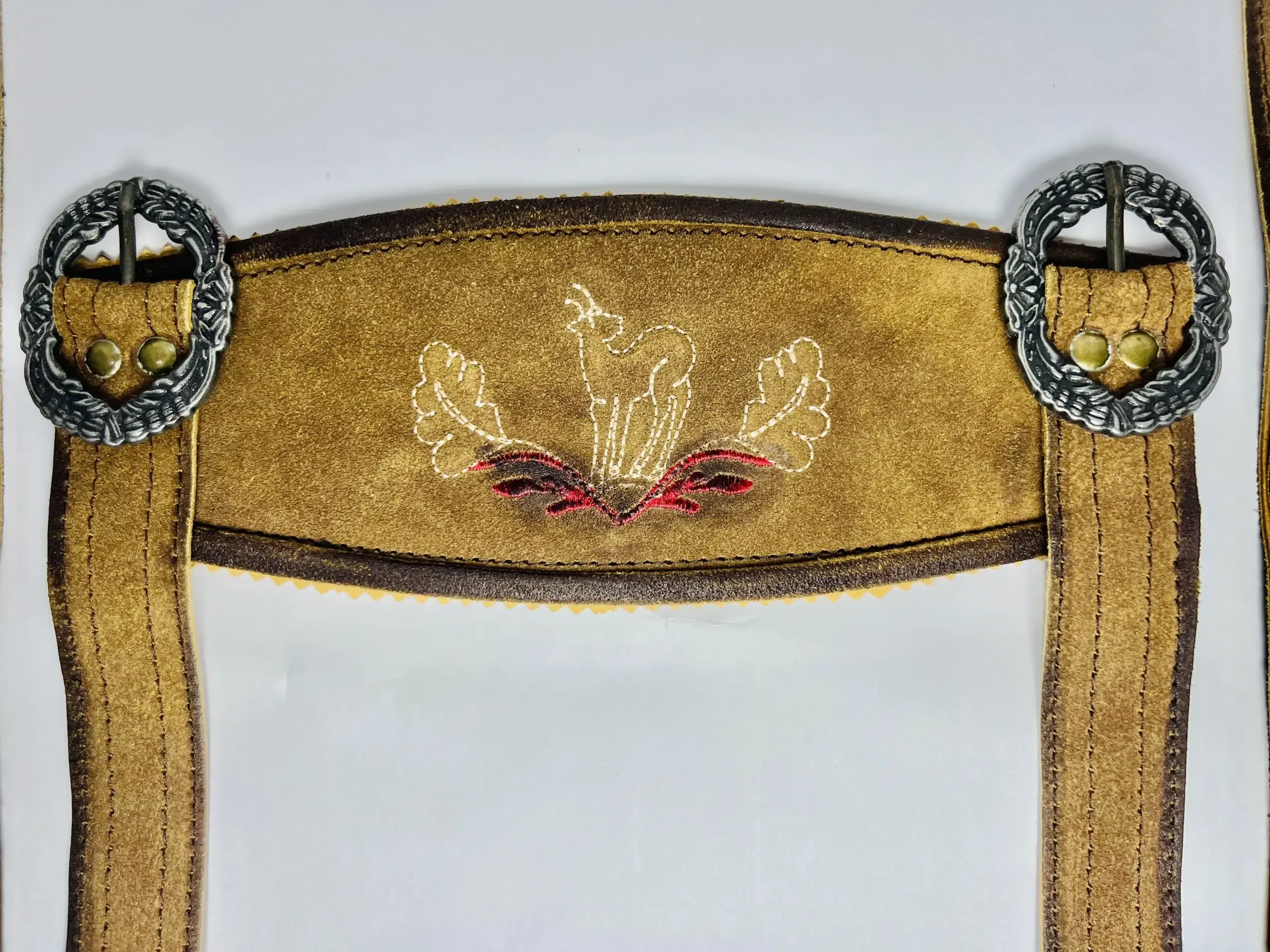 Close-up of a vintage brown Lederhosen suspender cross-bridge featuring white stag embroidery and ornate circular silver buckles. - Lederhosen For Sale